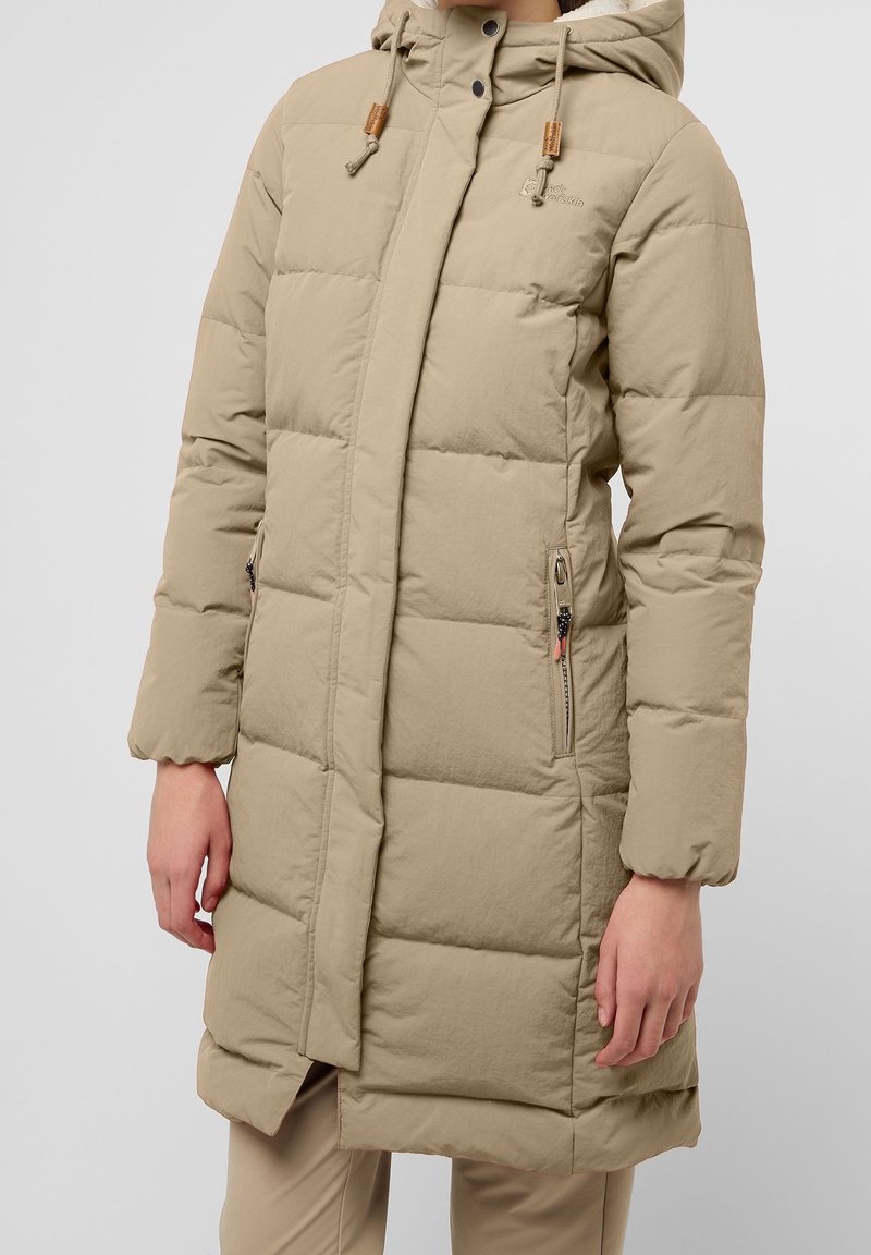 Beige vadderad jacka med huva, utrustad med tryckknappar och dragkedja, sidofickor och en förlängd design för värme och skydd.