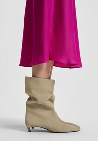 Khaki slouchy laarzen met een spitse neus en een lage hak, gemaakt van zachte suede, gecombineerd met een helderroze vloeiende jurk.