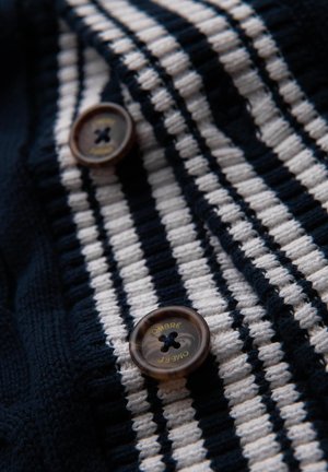Marineblau-weiß gestreifter Strickstoff mit einer strukturierten Oberfläche. Zwei runde, braune Knöpfe mit goldener "Ombre"-Textdetail.