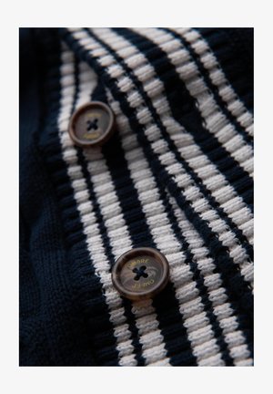 Tessuto a maglia a strisce blu navy e bianche con una finitura texture. Due bottoni rotondi marroni con dettagli in oro con la scritta "Ombre".