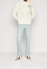 Hoodie crema con logo, jeans azzurri chiari e sneakers multi-color con dettagli in marrone, bianco e navy, vestibilità rilassata e texture morbida.