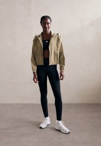 Kvinne som står innendørs iført en beige hettejakke med glidelås, svart sportsbh, svarte leggings, hvite sokker og hvite joggesko.