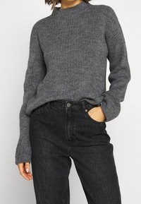 Femme portant un pull gris foncé à manches longues côtelé, rentré dans un jean noir taille haute, avec une main dans la poche.