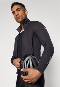 Veste de cyclisme noire avec col haut, design ajusté, logo Polartec® sur la manche. Le modèle tient un casque de vélo noir.