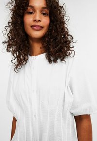 White button-up shirt with textured fabric, short puff sleeves, and a round neckline, featuring a subtle vertical ribbed pattern.