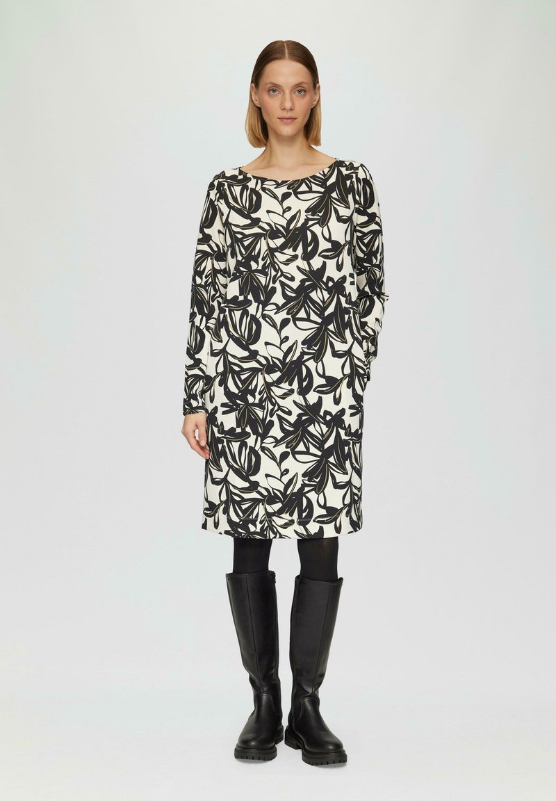 Robe florale noire et blanche avec des manches longues, un col rond et des poches, assortie à des bottes noires à hauteur de genoux. Tissu lisse avec un motif.