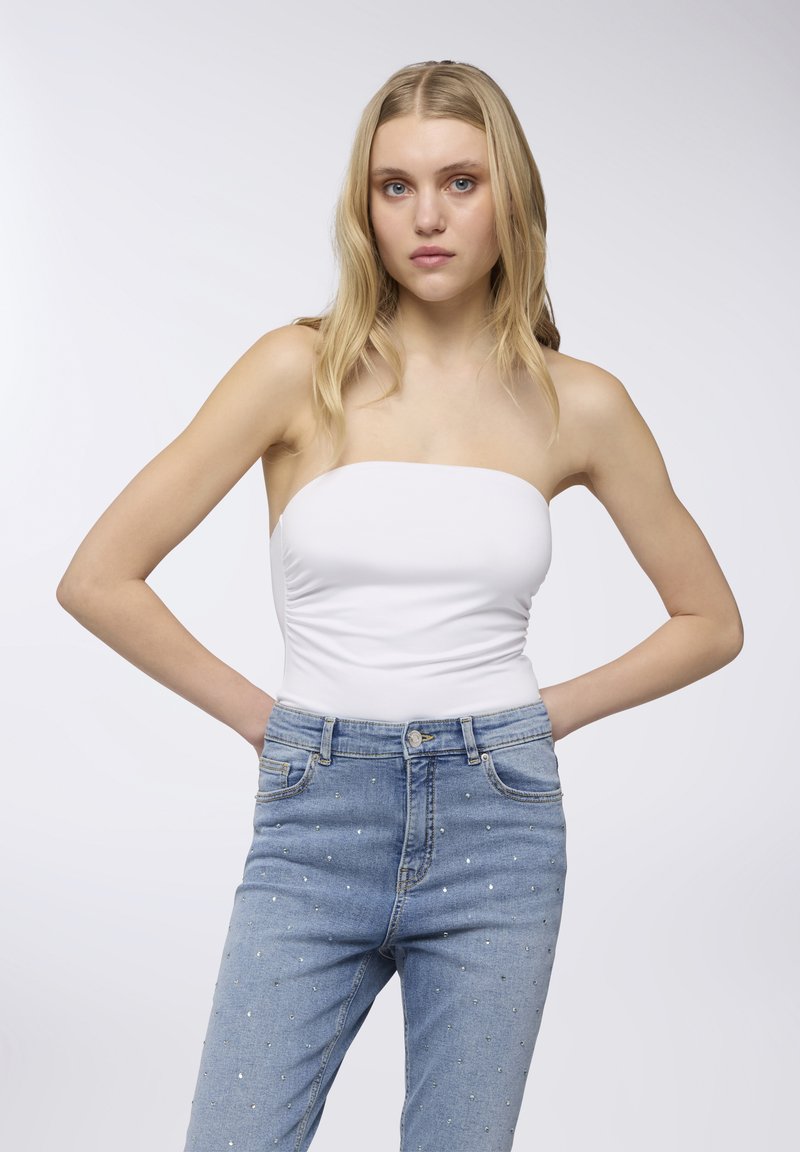 Top bianco senza spalline e jeans blu sbiaditi con borchie d'argento sparse. I jeans presentano un classico design a cinque tasche e una chiusura con zip.