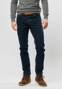 Jeans oscuros de corte slim con costuras en contraste, llevados con un suéter gris y botas de cuero marrón. Cinturón con hebilla de metal.