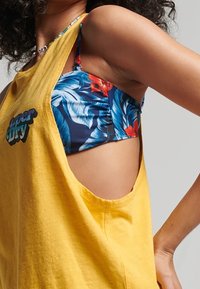 Top giallo senza maniche con un grande taglio laterale, caratterizzato da un logo nero. Sotto, è visibile un top bikini floreale blu.