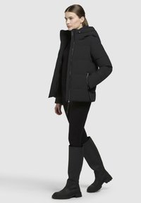 Doudoune noire avec capuche, fermeture éclair à l'avant et poches latérales. Portée par-dessus des leggings noirs et des bottes noires à hauteur de genou avec une semelle texturée.