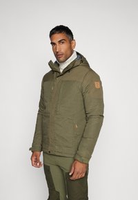 Groene gewatteerde jas met een capuchon, met ritssluiting zakken en een logo patch op de mouw. Gedragen over een geribbelde witte trui.