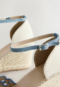 Sandales compensées beiges en tissu tissé avec brides de cheville en denim bleu et fermetures à boucle argentée, présentées en angle pour montrer la texture et les détails des coutures.