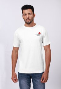 Λευκό βαμβακερό T-shirt με σχέδιο ενός κόκκινου ήλιου και μαύρου κύματος στην αριστερή περιοχή του στήθους. Στρογγυλή λαιμόκοψη και κοντά μανίκια. Απλός φόντος.