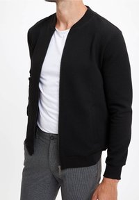 Czarna kurtka typu bomber z teksturowanej tkaniny, z prążkowanym kołnierzem i mankietami. Z przodu zapinana na suwak, noszona na białej koszulce i szarych spodniach.