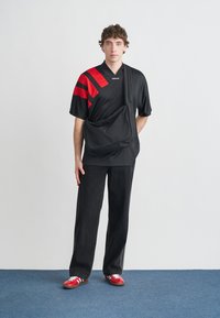 Čierny dresový top s červenými horizontálnymi pruhmi, skombinovaný s čiernymi širokými nohavicami a červenými teniskami. Má prívesok na tašku cez rameno.