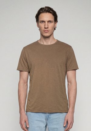 Giovane uomo con capelli corti castani che indossa una t-shirt marrone semplice e jeans azzurri chiari, in piedi davanti a uno sfondo grigio neutro.