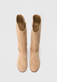 Beigen sävyiset mokkanahasta valmistetut polven korkeuteen ulottuvat saappaat, joissa on istuva varsi, pyöreä kärki ja kullanväriset vetoketjusomisteet sivulla. Pehmeä tekstuuri ja saumaton muotoilu.