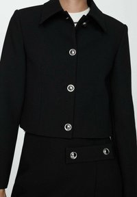 Sort cropped jakke med en skarp krave, sølv knaplukninger og en glat tekstur. Har en tætsiddende silhuet og et minimalistisk design.