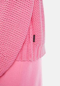 Rosa gestrickter Pullover mit einer groben Textur und einem gerippten Saum, versehen mit einem kleinen Logo-Tag. Kombiniert mit passenden, weichen rosa Sweatpants.