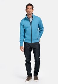 Blauwe ritsjas met hoge kraag, gemaakt van fleece-achtige stof; gecombineerd met donkere jeans en bruine suède schoenen.