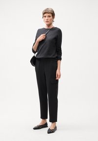 Crna dukserica, uske crne pantalone s prugama i ravne crne cipele s metalnim detaljima. Model drži malu crnu torbu, stojeći protiv bijele pozadine.