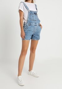 Jeansoverall med korta ben, ljusblå i färgen, med justerbara remmar, framfickor och en klassisk urblekt finish, bärs tillsammans med en vit t-shirt.