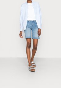 Camisa de botones azul claro sobre una camiseta blanca, combinada con pantalones cortos de mezclilla y sandalias plateadas. El conjunto presenta un ajuste relajado y un estilo casual.