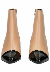 Botas de tobillo de cuero beige con forma cónica, que cuentan con una puntera negra brillante y cierre de cremallera en la parte posterior. Textura suave.