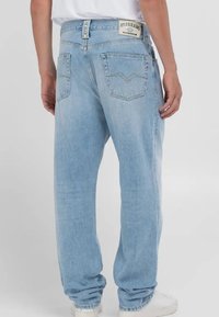 Bakifrån sedd person som bär ljusblå jeans med rakt ben och bakfickor, en vit skjorta och vita sneakers mot en enfärgad bakgrund.