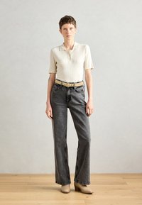 Polo shirt crème à côtes et manches courtes, associé à un jean noir taille haute et une ceinture beige. Modèle portant des bottes à talons carrés taupe.