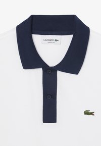 Camisa polo blanca con un cuello acanalado azul marino y tapeta de tres botones. Presenta un pequeño logo de cocodrilo verde en el lado izquierdo. Corte clásico.