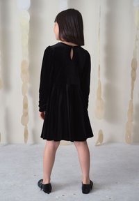 Kind steht mit dem Rücken zur Kamera, trägt ein schwarzes Samtkleid mit langen Ärmeln und schwarze Schuhe vor einem Hintergrund mit hängenden beigen Kreisen.