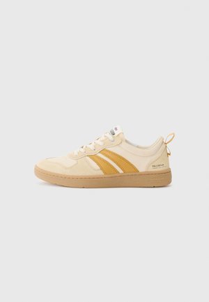 Beżowy sneakers o niskim profilu z żółtymi paskami po bokach, białymi sznurówkami, jasnobrązową podeszwą i uchwytem na pięcie, przedstawiony na jednolitym, jasnym tle.