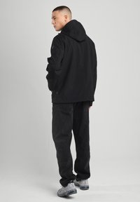 Black waterproof jacket with hood, featuring zipped side pockets and elastic cuffs, paired with black loose-fit pants and light sneakers.
