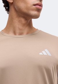 Mann trägt beige Rundhals-Shirt mit weißem Adidas-Logo auf der Brust, schaut zur Seite.
