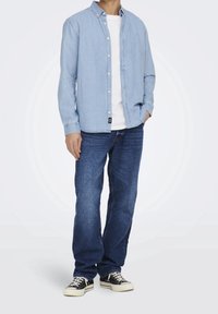 Camicetta di jeans azzurro chiaro sopra una maglietta bianca, abbinata a jeans blu scuro e sneaker nere. La camicia ha un colletto classico e bottoni frontali.