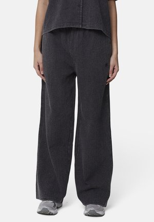 Persoon draagt losse, gestructureerde zwarte broek, bijpassende mouwloze top en grijze sneakers, staand op een witte achtergrond.