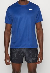 Man i en blå Nike-t-shirt och svarta mönstrade Nike-shorts, som står mot en enkel ljus bakgrund.