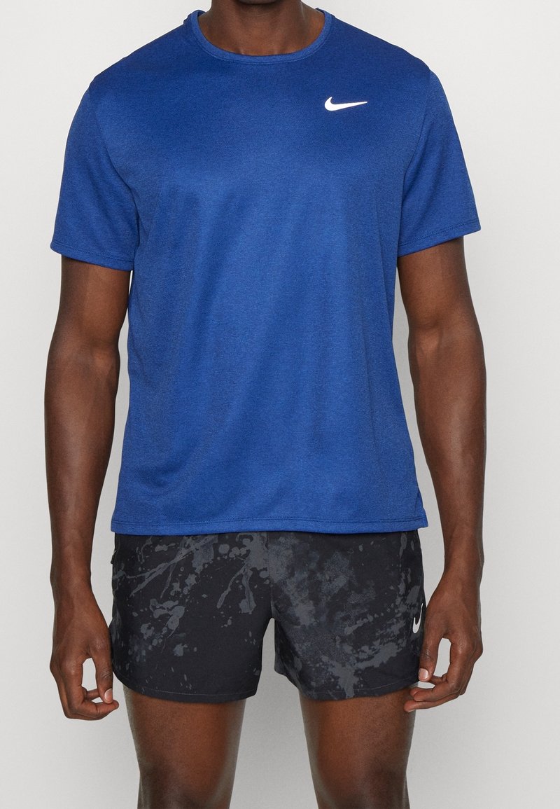 Man i en blå Nike-t-shirt och svarta mönstrade Nike-shorts, som står mot en enkel ljus bakgrund.