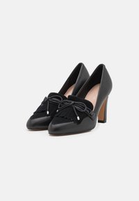 Scarpe nere con tacco alto, punta affusolata, dettagli in suede con frange e chiusura con lacci. Materiale in pelle liscia con un accento testurizzato.