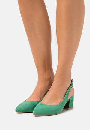 Femme portant des chaussures à talons blocs en suède vert avec une boucle en or, se tenant sur un fond blanc.
