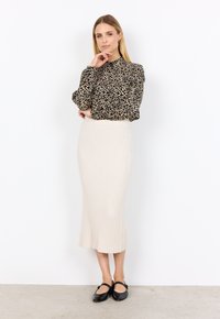 Schwarze und beige gemusterte Bluse mit langen Ärmeln und hohem Kragen, kombiniert mit einem figurbetonten, cremefarbenen, gerippten Midirock. Schwarze flache Schuhe runden den Look ab.