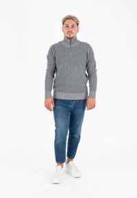 Maglione grigio con zip, con colletto e fondo a coste, abbinato a jeans tapered blu chiaro e sneakers bianche. Sfondo neutro.