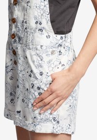 Rompers blanc à imprimé floral avec des motifs bleus et noirs, dotée de boutons en bois, de poches latérales et d'une section supérieure en tissu gris.