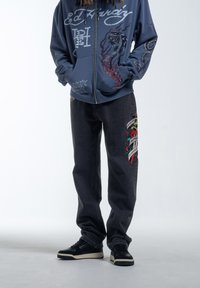 Marine blauwe hoodie met rits en grafische ontwerpen, gecombineerd met zwarte spijkerbroek met kleurrijke geborduurde patches en zwarte sneakers.
