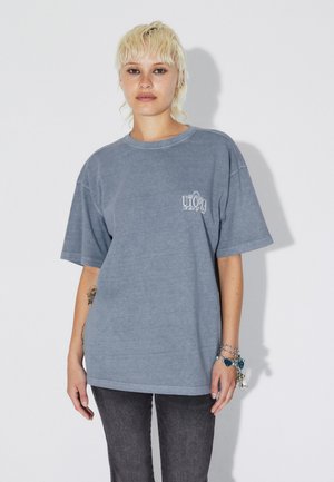 Camiseta gris extragrande hecha de tela suave, con un pequeño logotipo blanco en el pecho y mangas cortas. Combinada con jeans oscuros.