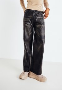 Black leather wide-leg pants with a slightly distressed texture, featuring front pockets and a high waist, styled with beige sneakers.
