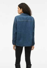 Giubbotto di jeans blu medio, con colletto classico, maniche lunghe e cuciture visibili. Abbinato a pantaloni neri aderenti.