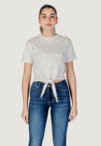 White cotton t-shirt with side cutouts and a front tie feature, adorned with beige embroidered logo design. Paired with blue denim jeans.