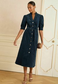 Abito midi blu navy con maniche a palloncino, scollatura a scallop, bottoni frontali e vita annodata. Indossato con sandali dorati e una borsa rotonda.